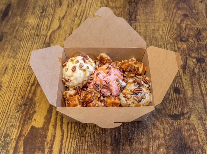 Three scoops of ice cream with caramel, pecans, and waffle pieces in a takeout box on a wooden table.