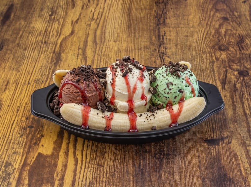 Banana split with chocolate, vanilla, and mint ice cream, cookie crumbles, and syrup on a wooden table.