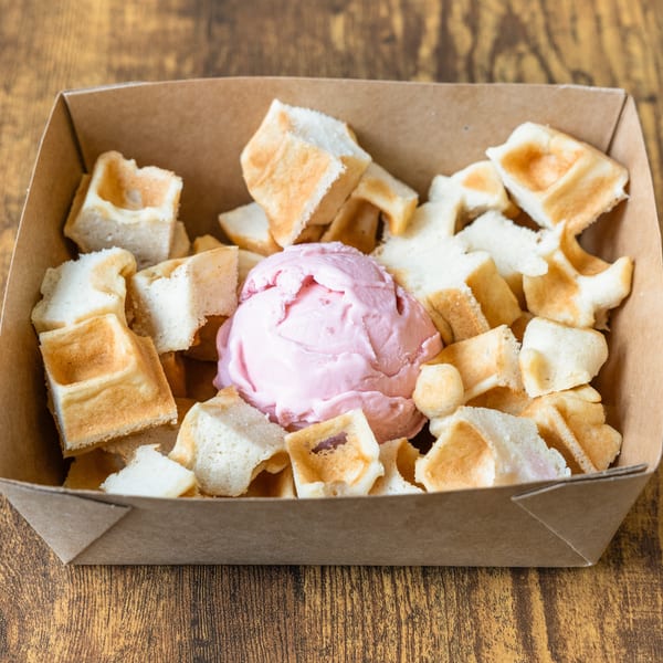 Waffle pieces and a scoop of pink ice cream in a brown cardboard container on a wooden table.