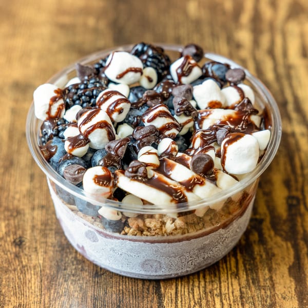 A dessert cup filled with blueberries, marshmallows, chocolate chips, and chocolate syrup on a wooden table.