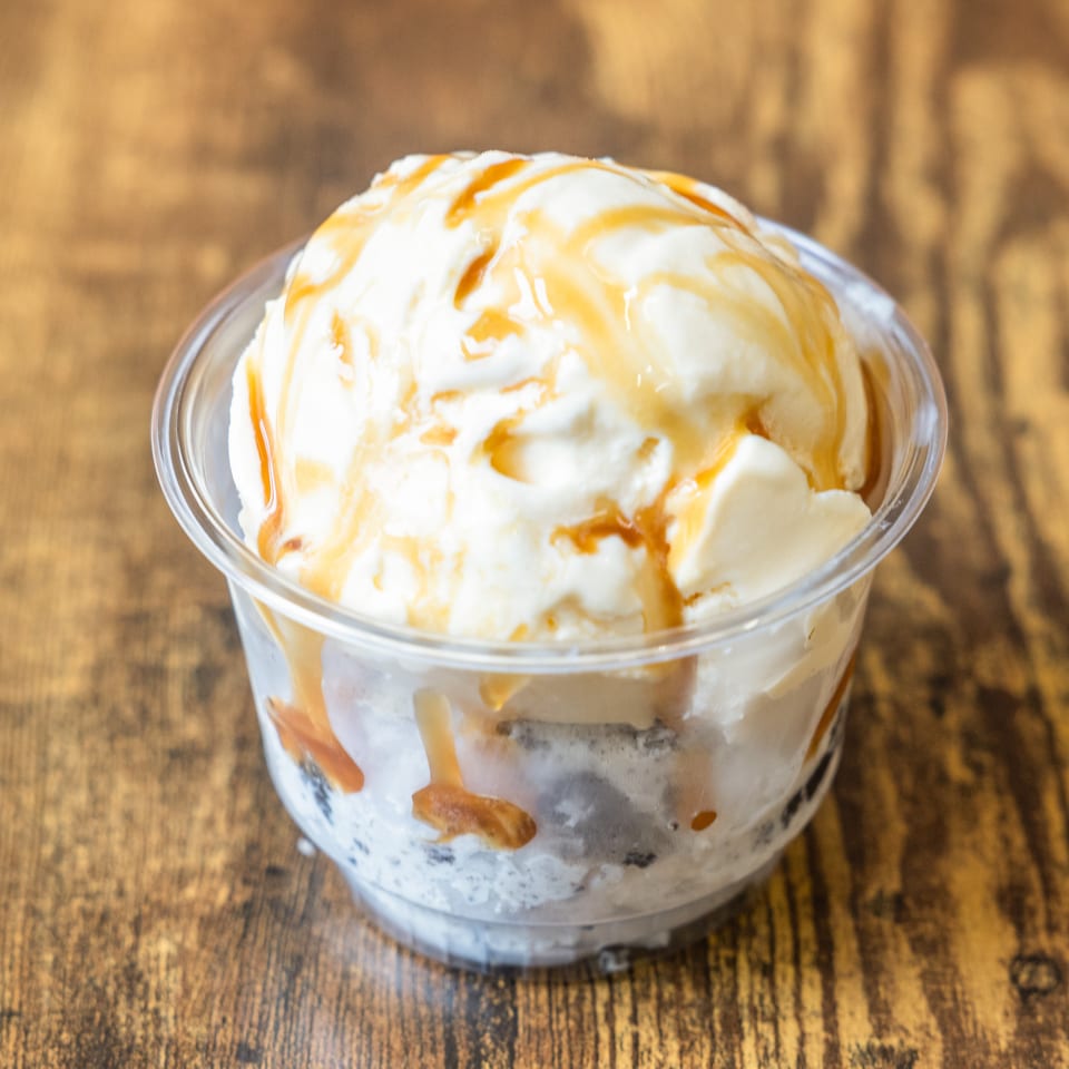 Vanilla ice cream with caramel drizzle in a clear plastic cup on a wooden table.
