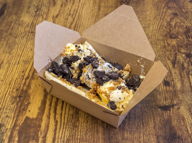 Vanilla ice cream with crushed cookies and granola in a brown takeout box on a wooden surface.