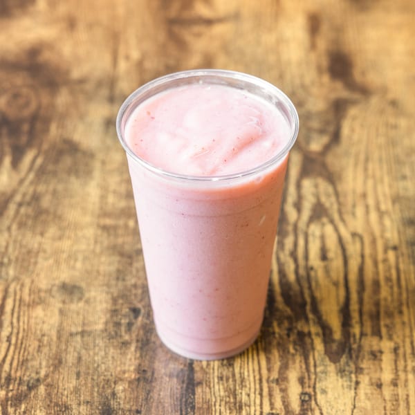 A tall, clear plastic cup filled with a light pink strawberry smoothie sits on a rustic wooden table.
