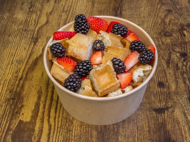 Waffle pieces with fresh strawberries and blackberries in a paper bowl on a wooden table.